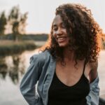 A joyful woman with curly hair smiles by a tranquil lakeside during sunset.