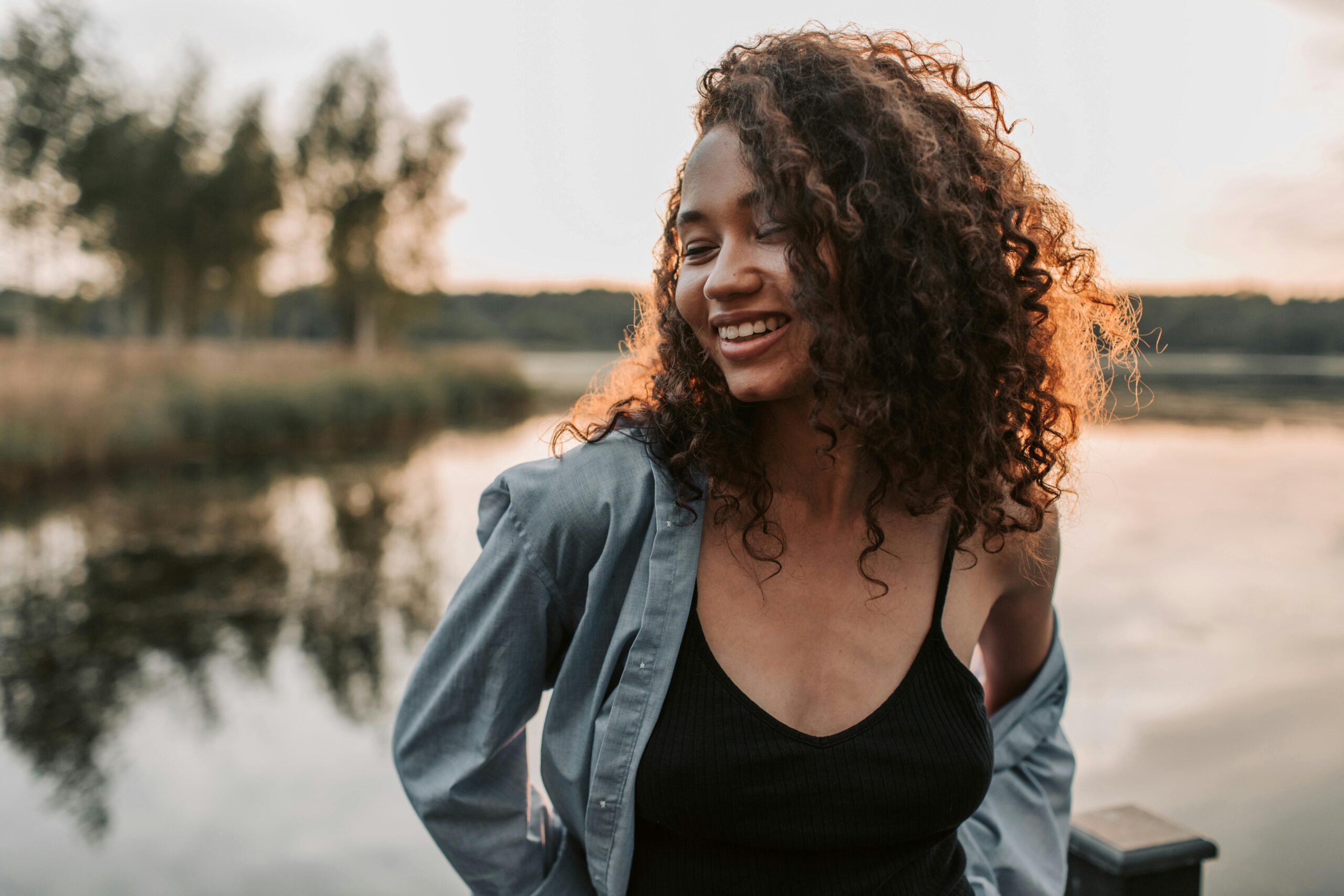 A joyful woman with curly hair smiles by a tranquil lakeside during sunset.