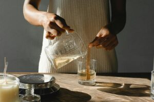 Unrecognizable African American female pouring melted shea butter from beaker into glass mold while making body butters at table